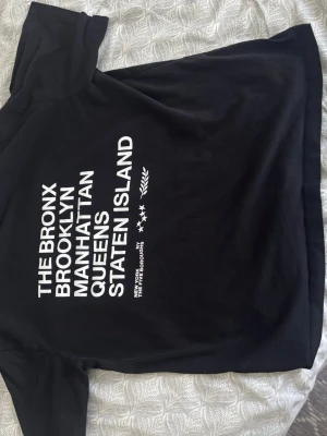 Svart t-shirt med New York-tryck - Cool svart t-shirt med vit text på ryggen där det står The Bronx, Brooklyn, Manhattan, Queens och Staten Island. T-shirten har klassisk passform och rund hals. Perfekt för dig som gillar streetstyle och vill ha en enkel men snygg look.