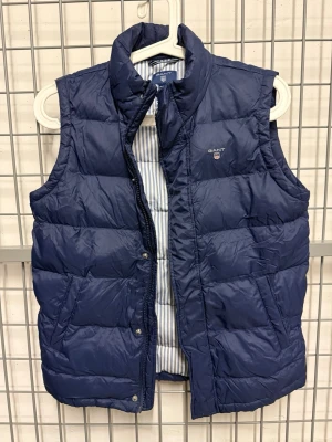 Mörkblå dunväst från GANT 146/152 - Stilren mörkblå dunväst från GANT med quiltad design och ståkrage. Västen har två fickor framtill och ett diskret GANT-logo broderat på bröstet. Insidan har ett randigt foder för extra detalj. Perfekt för lager-på-lager-stil.