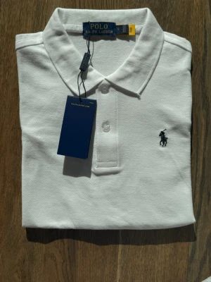 Vit pikétröja från Polo Ralph Lauren - Klassisk vit pikétröja från Polo Ralph Lauren med korta ärmar, krage och knappar framtill. Tröjan har den ikoniska broderade loggan på bröstet och är tillverkad i mjuk bomull. Perfekt till jeans eller shorts för en clean och stilren look. Riktig bra för golf vet inte om äkta 