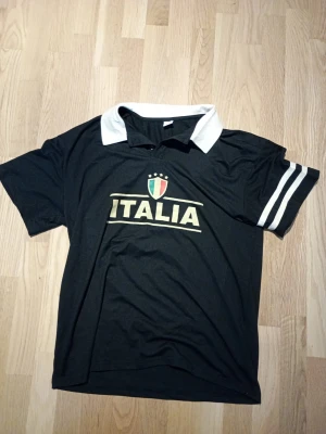 Svart Italia t-shirt med vit krage - Svart t-shirt med vit krage och två vita ränder på ena ärmen. Framsidan har ett stort 'ITALIA'-tryck och Italiens sköld. Tillverkad i 95% polyester och 5% spandex, perfekt för dig som gillar sportig stil.