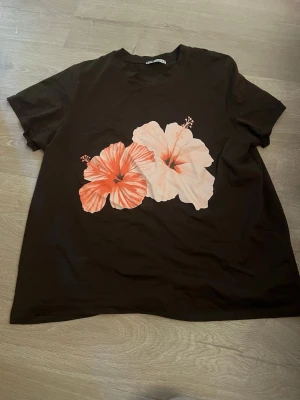 Svart t-shirt med blommor FB Sister - Svart t-shirt från FB Sister med två stora blommor i rosa och vitt framtill. Klassisk rund halsringning och korta ärmar. Perfekt för dig som gillar en chill och färgglad stil.