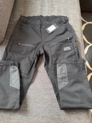 Mountain Trekking Friluftsbyxor - L/40, Short - Svarta byxor från Mountain Trekking med elastisk midja, flera fickor med dragkedja och förstärkta partier på knän och ben. Materialet är slitstarkt och designen är funktionell med smarta detaljer som passar för utomhusaktiviteter.                                                          Mått Från gren till fot: ca 73cm Midja: ca 41cm (lite stretch) Hott: ca 50cm