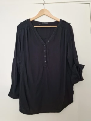 Svart blus med knappar från BPC collection - Stilren svart blus från BPC med v-ringning och knappar framtill. Blusen har långa ärmar som kan rullas upp och fästas med knapp, samt axeldetaljer. Tillverkad i ett lätt och luftigt tyg, perfekt för en avslappnad men snygg look. Storlek 40, fint skick. Från viskos.