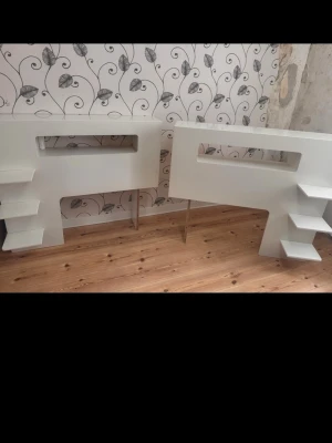 Vita hyllor med öppna fack - Två vita hyllor i modern design med öppna fack och hyllplan på sidorna. Hyllorna har rektangulära utskärningar i mitten och står på smala ben. Perfekta för förvaring eller dekoration i hemmet. Tillverkade i trä eller MDF med slät yta.