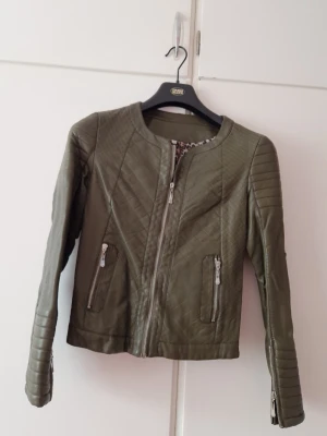 Olivgrön skinnjacka med dragkedjor - Snygg olivgrön skinnjacka med quiltade detaljer och raka dragkedjor framtill. Jackan har rund halsringning, två fickor med dragkedja och dragkedjor vid ärmsluten. Perfekt för en cool och stilren look.