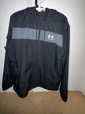 Svart vindjacka från Under Armour - Snygg svart vindjacka från Under Armour med dragkedja framtill och huva med dragsko. Jackan har en grå panel över bröstet med Under Armour-loggan tryckt i vitt. Tillverkad i lätt och tåligt material, perfekt för blåsiga dagar.