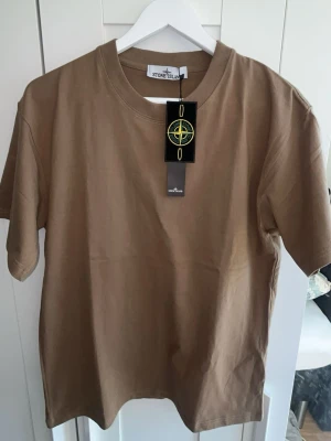 Beige t-shirt från Stone Island - Säljer en beige t-shirt från Stone Island med klassisk rund hals och korta ärmar. T-shirten är i mjuk bomull och har en enkel, clean design med logotypen på etiketten vid halsen. Perfekt för en avslappnad och stilren look.