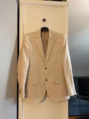 Beige linnékavaj med klassiska slag - Ljusbeige kavaj i linne med två knappar framtill, klassiska slag och tre fickor. Kavajen har rak passform. Perfekt för en stilren och avslappnad look. Använd 2 gånger.