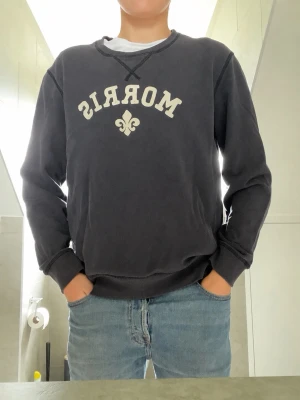 Morris mörkblå  - Mörkblå sweatshirt från Morris med vit logga och dekorativ detalj framtill. Klassisk rund halsringning och ribbade muddar vid ärmslut och nederkant. Perfekt till jeans för en avslappnad stil. 
