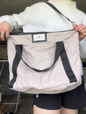 Beige shoppingväska från DAY - Stilren shoppingväska i beige/rosa nylon från DAY med svarta handtag och axelrem. Väskan har en rektangulär form, dekorativa blommönster på handtagen och en tydlig logotyp framtill. Perfekt för att bära med sig allt man behöver. Väskan passar till så många tillfällen, den har använts väl, går att platta ut!