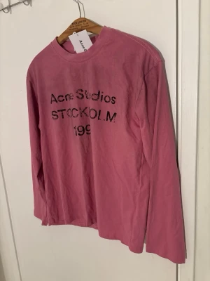 Rosa långärmad topp från Acne Studios - Cool rosa långärmad topp från Acne Studios med tryckt text 'Acne Studios STOCKHOLM' framtill. Tillverkad i mjuk bomull med relaxed fit och rund halsringning. Perfekt för en avslappnad och trendig look.