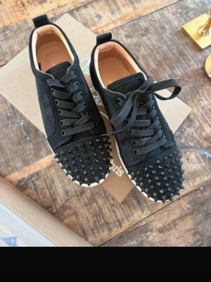 Svarta Louboutin sneakers med nitar - Svarta sneakers i mocka från Christian Louboutin med  svarta nitar på tån och klassisk röd sula. Skorna har svarta skosnören, låg profil och en vit kant runt sulan. Perfekta för dig som vill sticka ut med en edgy och lyxig stil. Säljer pga fel strl, tjejen jag köpte dem av saknade kvitto så osäker gällande äkthet. Har frågat ChatGPT som tyder på att skorna är äkta men som sagt ingen garanti. 