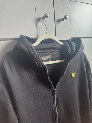Svart hoodie med dragkedja Lyle & Scott - Svart hoodie från Lyle & Scott med dragkedja framtill och klassisk huva. Bröstet pryds av den ikoniska gula loggan. Hoodien har två fickor framtill och är tillverkad i mjukt bomullsmaterial. Perfekt för en avslappnad och stilren look. XL i barnstorlek från kidsbrandstore(sitter som S)