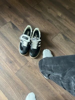 Adidas Wales Bonner svartvita sneakers - Svartvita Adidas Wales Bonner sneakers med klassiska vita ränder på sidorna, vit plös med Wales Bonner-detalj och beige sula. Skorna har en retro vibe och är tillverkade i en mix av mocka och läder. Perfekta för dig som gillar stilrena och unika sneakers.