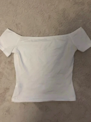 Vit offshoulder topp - Snygg vit offshoulder topp med korta ärmar och figurnära passform. Perfekt att styla med jeans eller kjol för en trendig look. Enkel och stilren design som passar till många outfits.