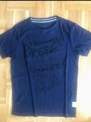 Mörkblå t-shirt med tryck - Snygg mörkblå t-shirt med svart texttryck på framsidan. T-shirten har rund halsringning med dubbla vita ränder på insidan av kragen och korta ärmar. Perfekt för en avslappnad och trendig stil.