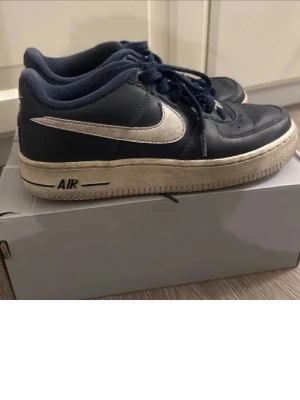 Nike Air Force 1 blå sneakers - Klassiska Nike Air Force 1 sneakers i blå läder med vit sula och vit swoosh-logga på sidan. Modellen har lågt skaft, perforerad tå och snörning framtill. Tidlös design som passar till många olika stilar.