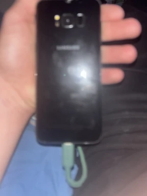 Svart Samsung Galaxy Android-mobil - Säljer en svart Samsung Galaxy med rundade kanter och glasbaksida. Telefonen har en kamera och fingeravtrycksläsare på baksidan samt Samsung-logga. Laddningskabel medföljer. Perfekt för dig som vill ha en stilren och pålitlig Android-telefon.