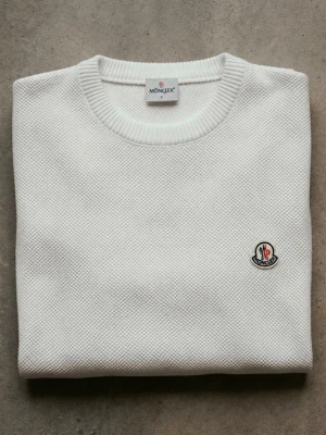 Vit stickad tröja från Moncler - Säljer en stilren vit stickad tröja från Moncler med rund halsringning och diskret Moncler-logga på bröstet. Tröjan har ribbade muddar vid ärmslut och nederkant samt en snygg struktur i materialet.