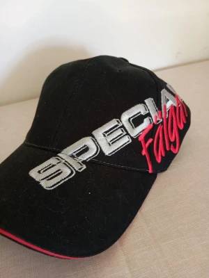 Svart keps med brodyr SPECIAL Falgas - Snygg svart keps med broderad text 'SPECIAL' i silver och 'Falgas' i rött på framsidan. Böjd skärm med röd detalj längs kanten. Perfekt accessoar för en cool och sportig stil.