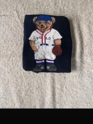 Mörkblå Polo Ralph Lauren björntröja - Stickad mörkblå tröja från Polo Ralph Lauren med ikonisk björn i basebolloutfit framtill. Tröjan har rund halsringning, ribbade muddar och är gjord i mjukt material. Perfekt för dig som gillar sportiga och lekfulla detaljer.