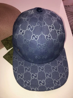 Blå keps från Gucci med GG-mönster - Snygg blå keps från Gucci med klassiskt GG-monogram över hela kepsen. Justerbar rem baktill och detaljer i läder. Tillverkad i canvasmaterial som ger en exklusiv känsla. Perfekt accessoar för att lyfta din stil.