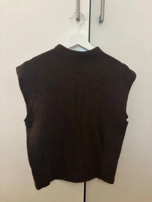 Mörkbrun stickad väst - En enkel och stilren mörkbrun stickad väst med hög hals. Västen har en rak passform och är perfekt att bära över en skjorta eller t-shirt för en avslappnad men snygg look. Aldrig använd!