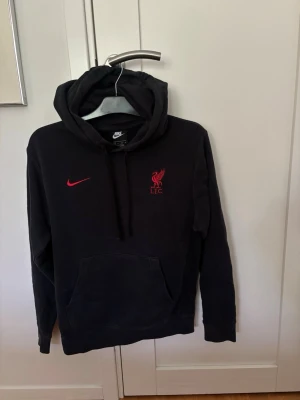 Svart Nike Liverpool hoodie - Snygg svart hoodie från Nike med Liverpool FC-logga och röd Nike swoosh på bröstet. Klassisk känguruficka framtill, dragsko i huvan och mjukt material. Perfekt för dig som vill visa ditt stöd för LFC med stil. Storlek XS, lite tvättblekt annars fint skick.