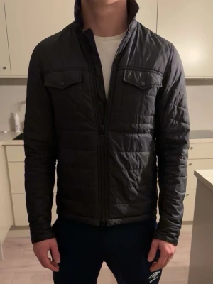 Svart pufferjacka med hög krage - Säljer en svart pufferjacka med hög krage och dragkedja framtill. Jackan har två bröstfickor med lock och en stilren, smal passform. Perfekt för kalla vinterdagar och ger en clean look till din outfit.