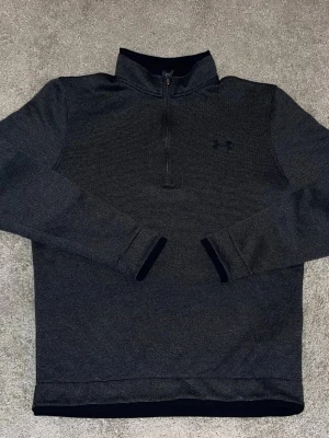 Mörkgrå halvzip från Under Armour - Snygg mörkgrå långärmad tröja från Under Armour med halv dragkedja och diskret logga på bröstet. Tröjan har ståkrage och är tillverkad i ett mjukt, strukturerat material som känns sportigt och bekvämt.