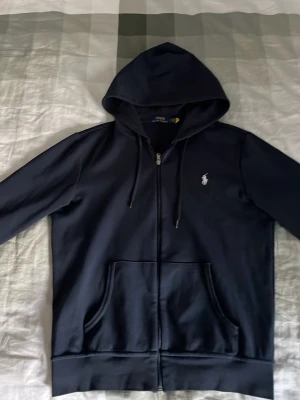 Svart hoodie från Polo Ralph Lauren - Snygg svart hoodie med dragkedja från Polo Ralph Lauren. Klassisk design med huva och dragsnören, två stora fickor framtill och den ikoniska vita loggan broderad på bröstet. Perfekt för en avslappnad och stilren look.