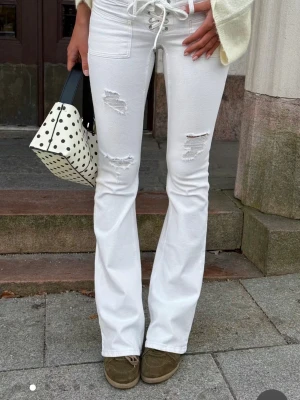 Vita bootcut jeans med snörning - Snygga vita bootcut jeans med snörning i midjan och slitna detaljer på låren. Jeansen har hög midja och utsvängda ben, vilket ger en trendig siluett. Perfekta för dig som vill sticka ut med en cool och modern look.