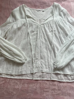 Vit blus från Dobber - Luftig vit blus från Dobber med v-ringning och långa ärmar. Blusen kan knytas framtill och är tillverkad i ett lätt, strukturerat tyg som ger en avslappnad boho-känsla. Perfekt för sommaren. Storlek L men passar som en storlek M