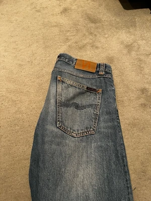 Blå jeans från Nudie Jeans - Säljer ett par klassiska blå jeans från Nudie Jeans med snygg tvätt och kontrastsömmar. Jeansen har raka ben, två bakfickor med den ikoniska vågstickningen och patch i läder bak i midjan. Perfekta för en avslappnad och trendig stil. Jeansen är i jättebra skick och säljs pga att de är för små. 