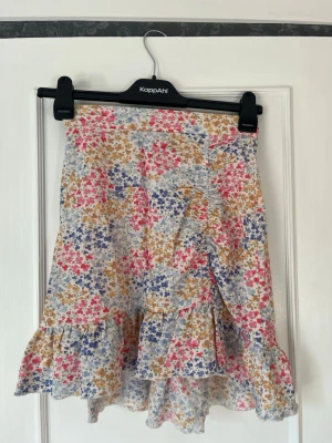 Blommig volangkjol från Gina Tricot - Supersöt kort kjol från Gina Tricot med blommigt mönster i rosa, blått och gult. Kjolen har volang nertill och en lätt omlottkänsla framtill. Perfekt för varma dagar och ger en fräsch och somrig vibe. I storlek XS❣️ 