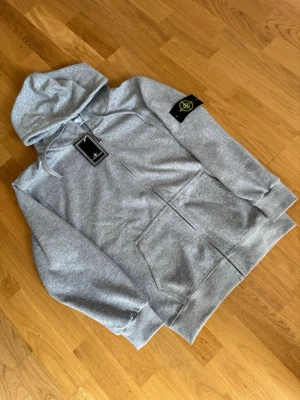 Grå hoodie från Stone Island - Snygg grå hoodie från Stone Island med dragkedja framtill, klassisk huva med dragsko och känguruficka. Ikonisk Stone Island-patch på vänster ärm. Tillverkad i mjuk bomullsblandning som känns skön mot huden. Perfekt för en avslappnad streetwear-look.