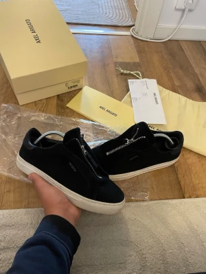 Svarta sneakers Axel Arigato med dragkedja - Snygga svarta sneakers från Axel Arigato i mocka med unik dragkedja framtill istället för klassiska snören. Skorna har vit sula och minimalistisk design med diskret logga på sidan. Perfekta för dig som gillar stilrena och moderna detaljer. Storlek 41 passar 42