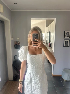 Vit kort klänning med puffärmar - Superfin vit kort klänning med puffiga kortärmar och fyrkantig urringning. Klänningen har ett subtilt blommigt mönster i tyget som ger en lyxig känsla. Perfekt för sommarens alla tillfällen och har en figurnära passform. Passar perfekt för studenten eller avslutning!
