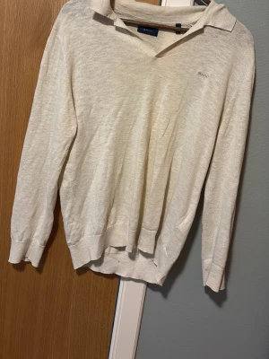 Beige stickad tröja från GANT - Klassisk beige stickad tröja från GANT med krage och v-ringning. Tröjan har långa ärmar och ribbade muddar