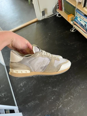 Valentino beiga sneakers med nitar - Snygga sneakers från Valentino i beige och grå toner med camomönster, mesh och mockadetaljer. Skorna har coola nitar på hälen och en chunky sula. Perfekta för dig som gillar streetstyle och exklusiva märken.