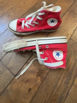 Röda Converse All Star Highs - Klassiska röda Converse All Star med högt skaft och vita snören. Skorna har vit gummisula och tå, samt den ikoniska loggan på sidan. Perfekta för en cool och avslappnad stil.