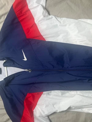 Nike vindjacka blå röd vit - Snygg vindjacka från Nike i blått, rött och vitt med dragkedja framtill och klassisk vit Nike-logga på bröstet. Jackan har sportig design och är tillverkad i lätt,  material. Perfekt för en streetwear-look. Dragkedjan är lite svår ibland men funkar som den ska. 
