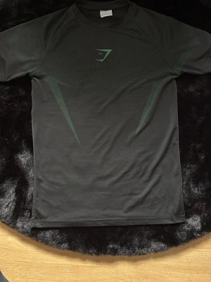 Svart träningströja från Gymshark - Svart kortärmad träningströja från Gymshark med diskreta gröna detaljer och logga på bröstet. Tillverkad i ett stretchigt och fukttransporterande material som passar perfekt för intensiva träningspass.