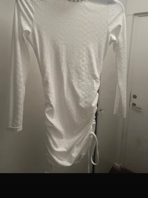 Vit långärmad klänning med snörning - Snygg vit klänning med lång ärm och diskret strukturmönster. Klänningen har snörning i sidan som ger en cool draperad effekt. Perfekt för dig som gillar stilrena och trendiga plagg.