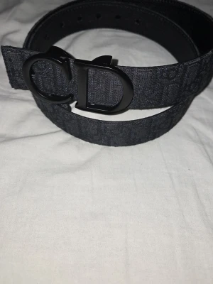 Svårt zara bälte med CD-spänne - Snyggt svart bälte från Zara med stort CD-spänne i metall. Bältet har diskret mönster längs hela bandet och är tillverkat i textilmaterial med detaljer i skinn. Perfekt accessoar för att lyfta din outfit.