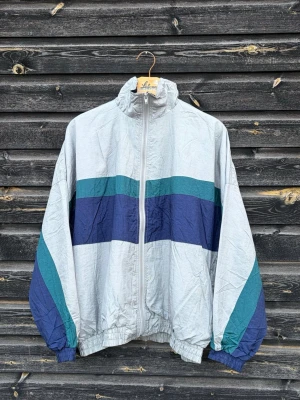 Retro vindjacka med blåa ränder - Snygg retro vindjacka i ljusgrått med breda blå och turkosa ränder över bröstet och ärmarna. Jackan har dragkedja framtill, hög krage och resår vid ärmslut och midja. Perfekt för en sportig och avslappnad stil.