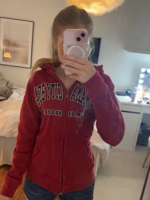 Röd hoodie med dragkedja och tryck - Röd hoodie med dragkedja, huva och fickor framtill. Tryck på bröstet med texten 'CANADA CITY BEAR'.OBS! Tröjan är gammal och trycket smular sig/lossnar lätt. Säljes därför billigt i detta skick. Tyget är i övrigt helt.