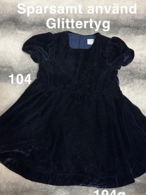 Mörkblå glittrig klänning med puffärm - Mörkblå klänning i glittrigt tyg med puffärmar och rund halsringning. Klänningen har en utställd kjol och är perfekt för dig som gillar lite extra glitter. Materialet känns mjukt och har en lyxig känsla.🌸 Sparsamt använd. Glittertyg. 104