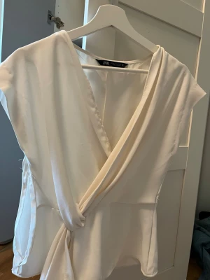 Vit omlottblus från Zara - Stilren vit omlottblus från Zara med knytning framtill och djup v-ringning. Blusen har korta ärmar och är tillverkad i ett mjukt, lätt material som ger en snygg och avslappnad look.