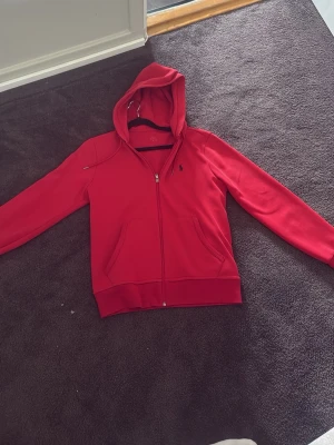 Röd hoodie från Polo Ralph Lauren - Snygg röd hoodie från Polo Ralph Lauren med dragkedja och huva. Klassisk logga broderad på bröstet och två fickor framtill. Mjukt material och ribbade muddar vid ärmslut och nederkant. Perfekt för en avslappnad stil.